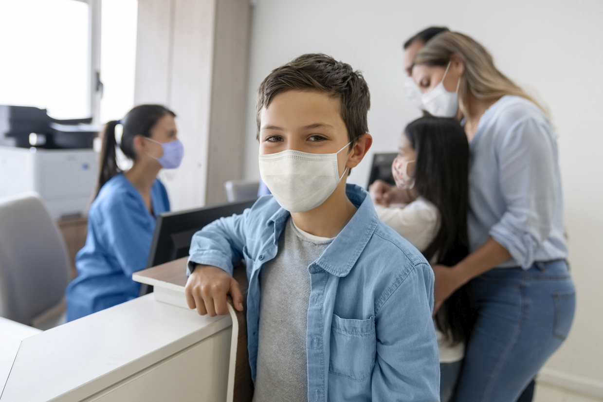Junge mit medizinischem Mund-Nasen-Schutz steht am Tresen in einer Arztpraxis