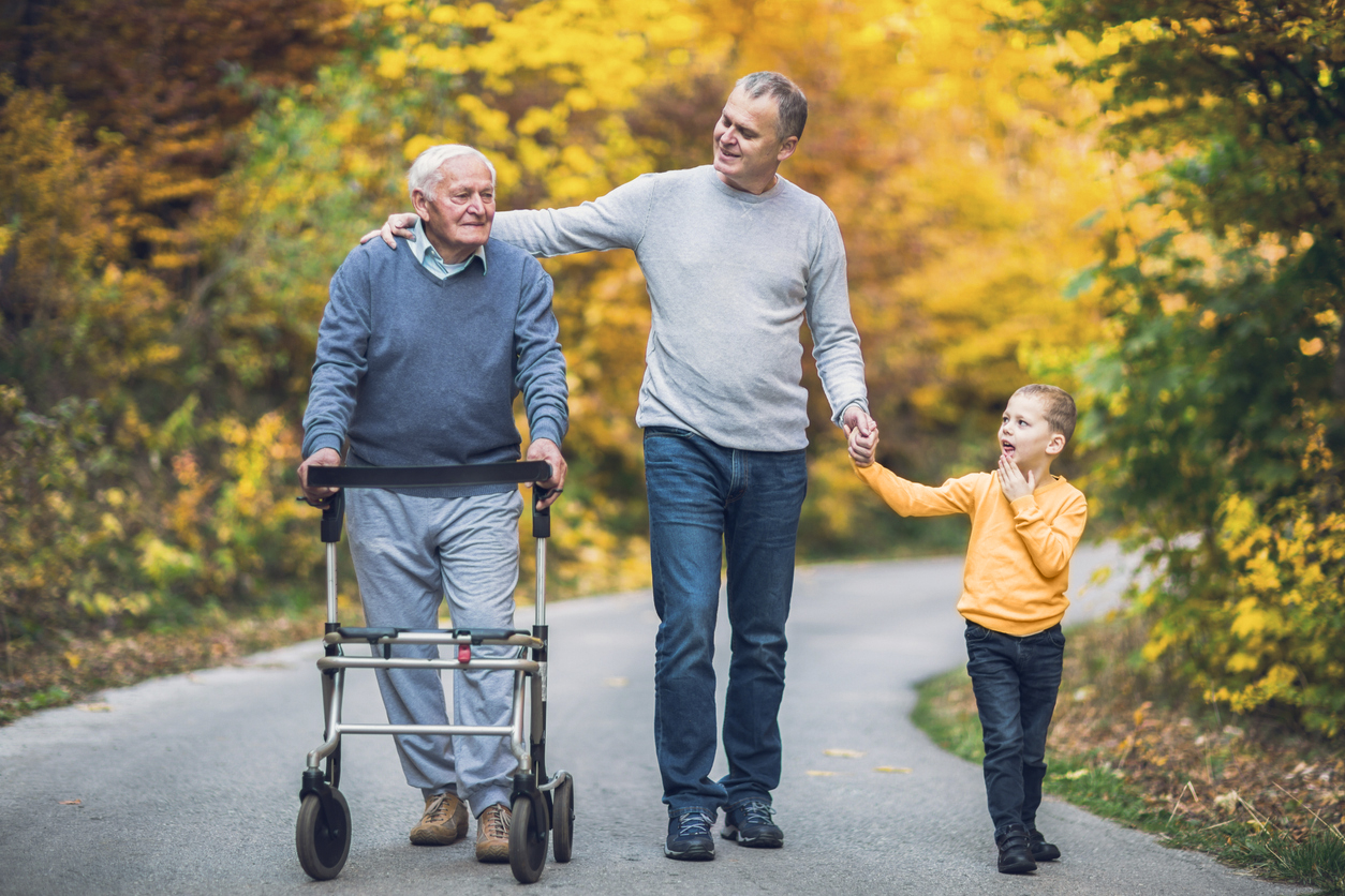 Drei Personen aus drei Generationen gehen gemeinsam spazieren, die älteste Person schiebt einen Rollator