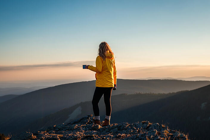 Frau mit gelber Jacke genießt auf dem Berggipfel die Aussicht mit einem Getränk.