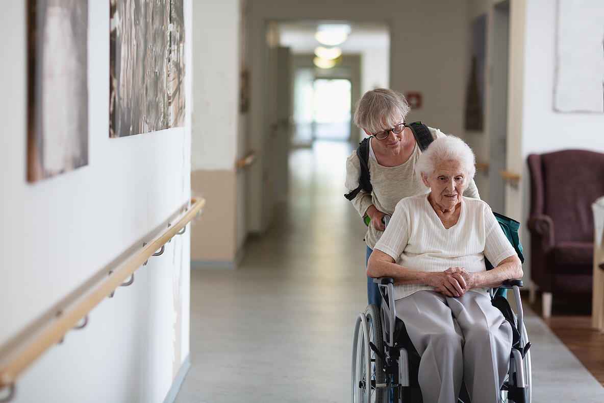 Alte Dame wird im Rollstuhl über einen Flur geschoben